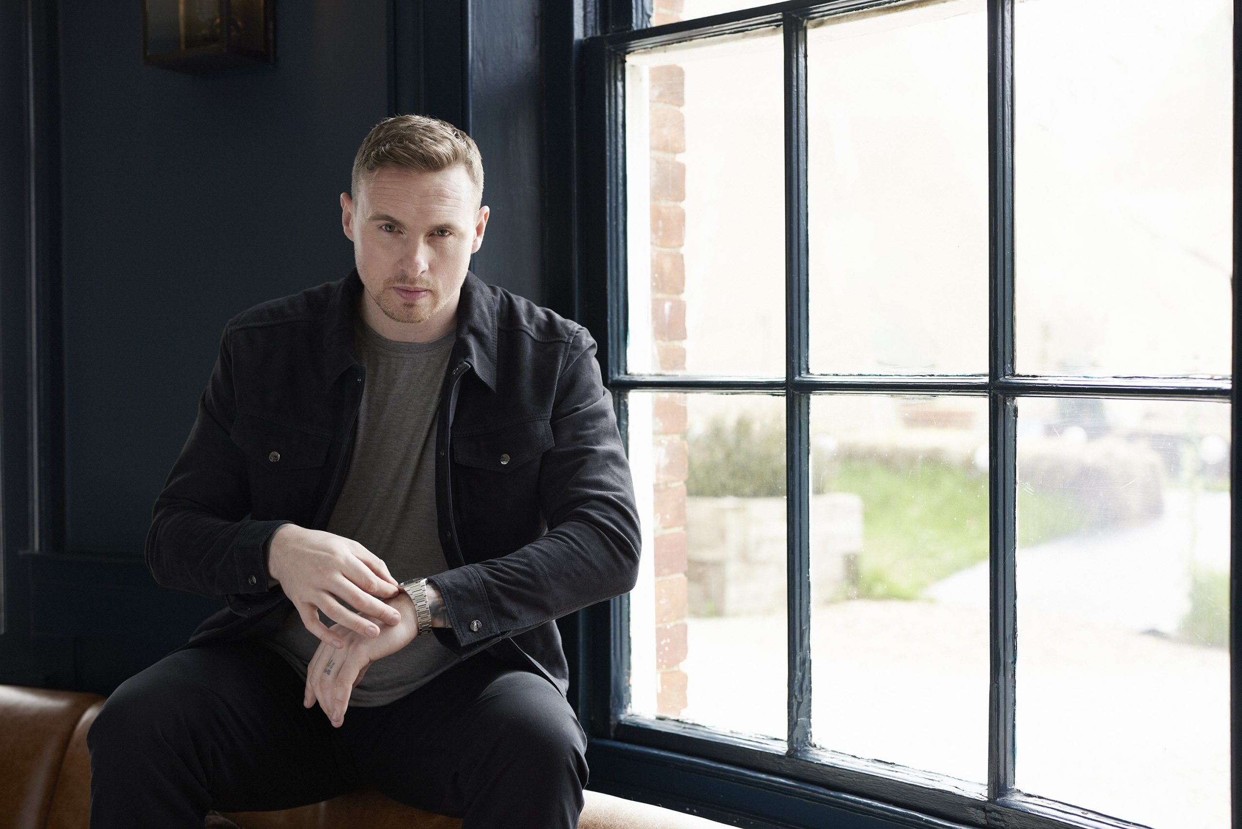 David Stockdale checking his watch while sat on a leather sofa in front of a large window