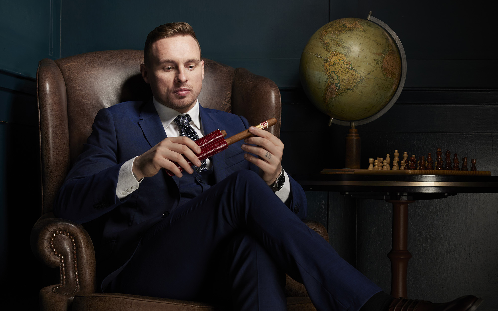 David Stockdale sat in a leather armchair wearing a blue suit holding a cigar